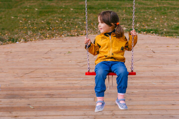 Happy little girl swinging on a swing in the Park in beautiful autumn, or cold summer. Cute child 3-4 years in a jacket and jeans having fun on a playground outdoors. Adorable girl in a park.