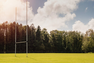 Goal post for Irish national sports rugby, hurling, caomogie, football by trees on a sunny warm day. Nobody, Cloudy sky, Sun flare, Shadow on grass.