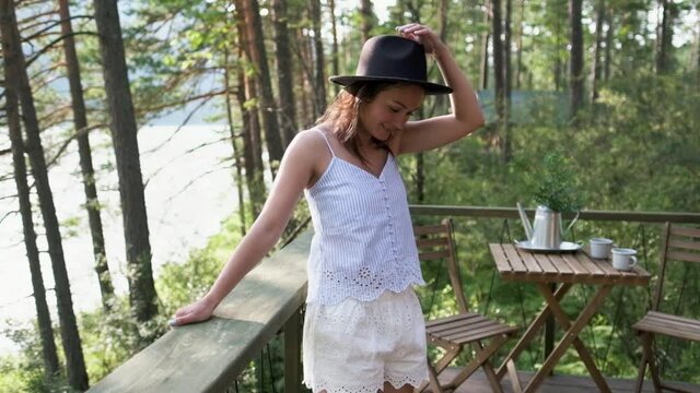 young woman in shorts, a t-shirt and a black hat enjoys nature on a terrace with mountain views. Attractive brunette covers her face with a hat enjoys nature in the mountains