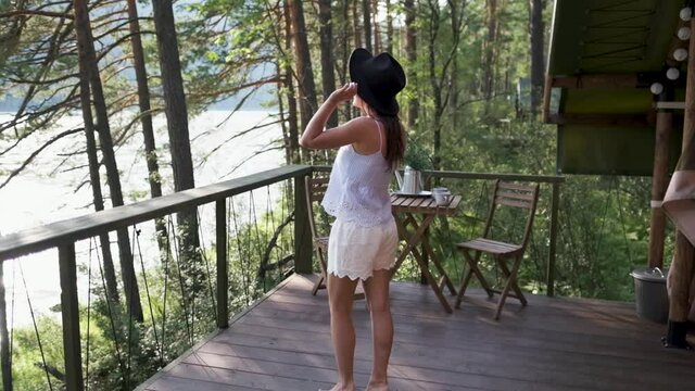 young woman in shorts, a t-shirt and a black hat dancing on a terrace with mountain views. Attractive brunette covers her face with a hat enjoys nature in the mountains
