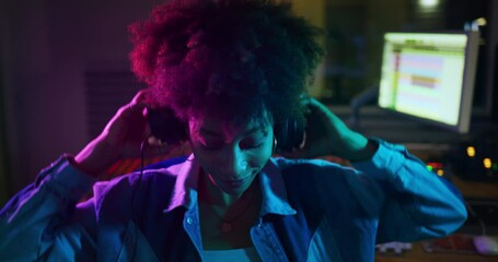 An young happy professional african female producer is taking off headphones while recording a new song and smiling satisfied in camera in a music studio with colorful lights on a background. - Powered by Adobe
