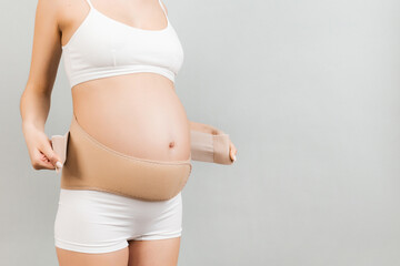 Close up of pregnant woman in underwear dressing elastic maternity band at gray background with...