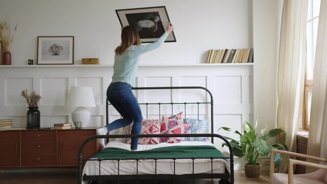 young woman tries on picture to new place in bedroom in new apartments, puts it on shelf and sits on bed, looks on it and admires. finishes relocation and cozy interior design.