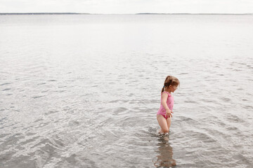 Fototapeta premium A three-year-old girl of European appearance plays in the water on the beach. A child walks ankle-deep in the lake. Holidays in nature. Summer photo of a baby in a swimsuit.