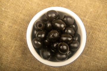 Black seedless olives in a ceramic bowl on a background of coarse-textured fabric. Close up.