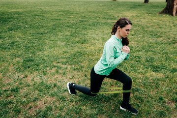 Side view of attractive sportswoman doing lunges with resistance band in park