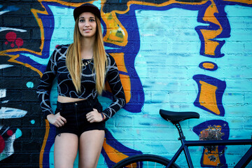 Portrait of pretty hipster girl posing on graffiti wall background outdoors, young woman standing with her bike on colorful wall background