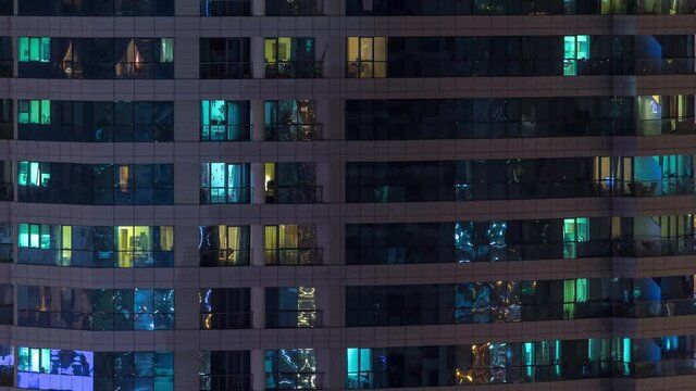 Rows of glowing windows with people in the interior of apartment building at night. Modern skyscraper with glass surface. Concept for business and modern life. Tilt down