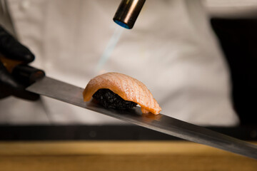 Chef making traditional sushi nigiri with black rice. Close up of preparing salmon Tataki. Process of Roasting salmon on Japanese knife with fire. Japanese food in restaurant. Cooking Asian dish