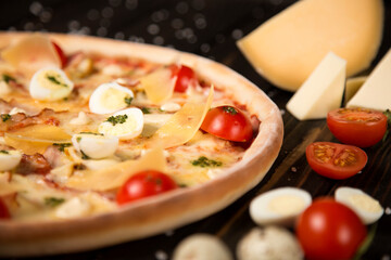 Side view of pizza with cherry tomatoes, pesto sauce, parmesan, cheese, chicken fillet, bacon, quail eggs, mozzarella cheese on wooden background with sea salt and ingredients. Close up of uncut pizza
