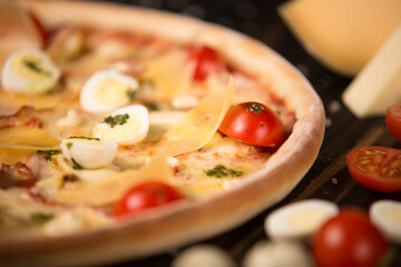 Close up of pizza with cherry tomatoes, pesto sauce, parmesan, cheese, chicken fillet, bacon, quail eggs, mozzarella cheese on wooden background with sea salt and ingredients. Half of uncut pizza
