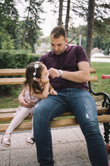 Man pinching little girl's cheek while resting on bench. Father and his little daughter sitting on wooden bench in city park and playing.