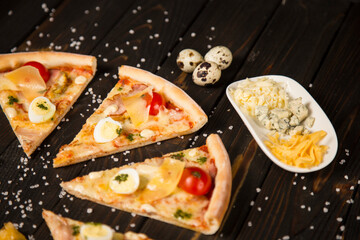 Slices of pizza with tomatoes, pesto sauce, bacon, quail eggs, mozzarella and Parmesan cheese on wooden background with sea salt and ingredients. Cheese plate and quail eggs near pizza pieces