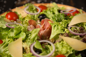 Close up of Traditional Italian pizza with jamon Serrano in focus, Parmesan cheese, black olives and cherry tomatoes on wooden background. Tasty Mediterranean rustic meal. fast food delicious dish
