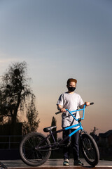 Fototapeta premium Portrait of a teenager in the sunset in a black mask on a blue bicycle. Quarantine. It is forbidden to ride a bicycle. Covid-19.