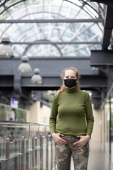 Portrait of a beautiful woman in a black mask, which stands inside a building with beautiful architecture. Woman in a dark green sweater and khaki pants.