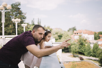 Father and daughter leaning on fence and looking at view. Man hand pointing while hugging his little daughter on fence outdoor.
