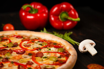Side view of pizza with bell pepper, green olives, smoked sausages, mozzarella cheese, pepperoni, salami, chili pepper, champignons on wooden board with ingredients on background. uncut tasty pizza
