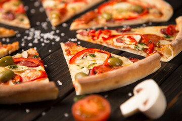 Close up of sliced pizza with bell pepper, olives, sausages, mozzarella cheese, pepperoni, salami, chili pepper on wooden board with sea salt, tomato and champion on background. Rustic food
