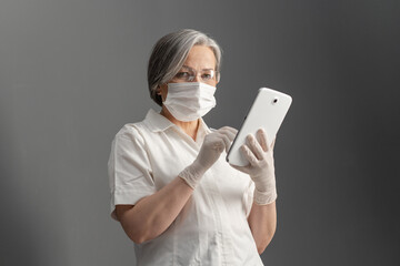 Fototapeta na wymiar Serious female doctor works with tablet computer. Caucasian woman in white protective uniform uses digital tablet looking at camera while standing on gray wall background.
