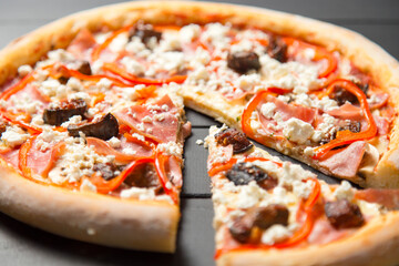 Close up of traditional Italian pizza with ham, beef, bell pepper, mozzarella cheese and tomato sauce on dark wooden background. Slice of pizza moved near the whole dish. Delicious Mediterranean meal