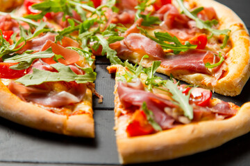 Close up of traditional Italian pizza with jamon, arugula and cherry tomatoes on dark wooden background. Slice of pizza moved near the whole dish. Delicious Mediterranean meal with fresh herbs