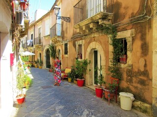 Italy, Sicily, Siracusa, Ortigia - 2018 August 17th - Traditional narrow street in Ortigia, Syracuse; woman with flower colorful dress strolling