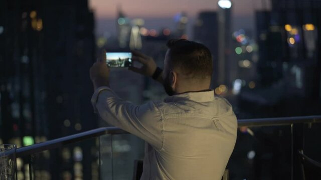 Young man photo of city skyline with smartphone on terrace at night