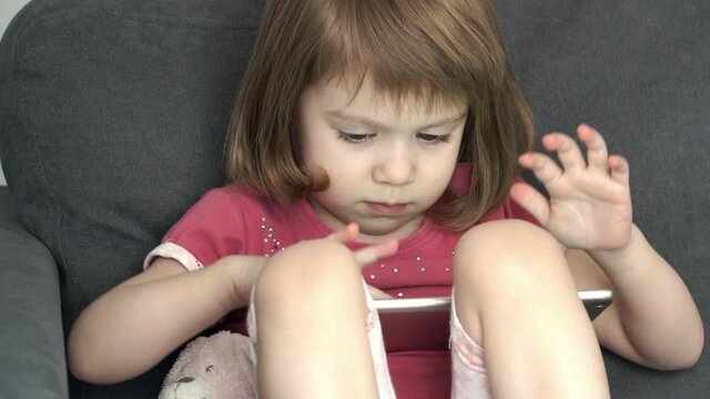 Portrait of little serious 3 year old girl attentively looks at digital tablet, enthusiastically touches fingers of screen and studies Internet
