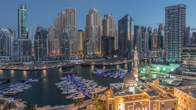 Yachts in Dubai Marina flanked by the Al Rahim Mosque and residential towers and skyscrapers aerial night to day transition timelapse. Modern high rise skyline with boats and wooden dhows