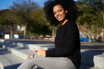Fototapeta premium Cheerful young attractive female dressed in casual wear holding smartphone look into camera while sitting outdoors, gorgeous afro american hipster girl recreating enjoying sunny weather on weekends