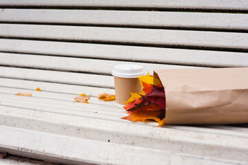 A brown paper cup for coffee with a white plastic lid and a paper bag full of red and yellow maple leaves lie on an old scarlet white bench on a cloudy autumn day.