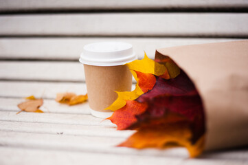 A paper cup with a white lid stands on a white old bench next to a paper bag, inside which are colored leaves of maple.