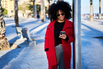 Cheerful trendy dressed afro american female tourist strolling having city tour using navigation on smartphone searching right direction to showplaces enjoying sunny weather and weekends free time