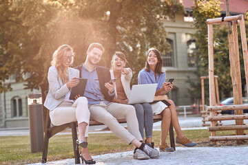 group of young caucasian businesspeople laughing at bench in a park with their cell phones, laptop, tablet. Looking at camera, eye contact. Break, pause from work.