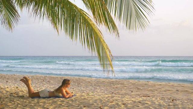 Young Caucasian woman lies on the tropical white sand beach and watches the surfers catching waves. Tourist girl relaxes on the scenic coastline of Barbados and observes the approaching ocean waves.