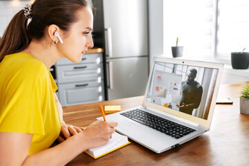 An African-American male teacher conducts webinar, online classes on the laptop screen indoor, young woman is watching and taking notes in the kitchen at home. Online studying concept