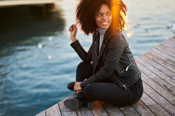 Pondering happy gorgeous woman dressed in trendy clothing looking away during resting on wooden pier near to lake at sunset time.Smiling young attractive hipster girl having recreation outdoors