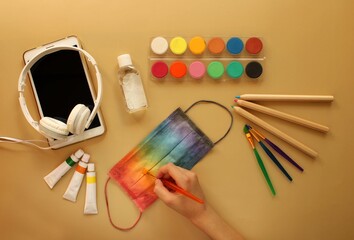 A child's hand paints a protective mask in rainbow colors next to school stationery, antiseptic, electronic tablet and headphones. Yellow background. Back to school and protection against coronavirus