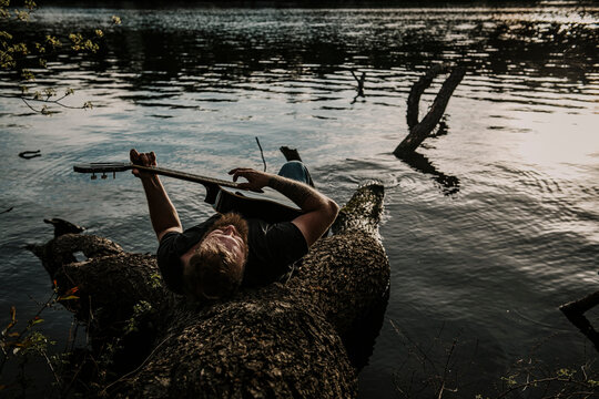 Europe, Germany, Berlin, Mueggelheim, Grosse Krampe, man relaxing on a tree in the lake with his guitar.