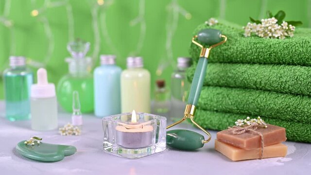green jade roller leaned on towels between candle and soap bars against blurry facial serum bottles in spa salon closeup
