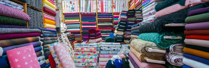 clothes in shop, Rolls of fabric and textiles for sale stacked on shelves in shop, View of cloth rolls of different colors and patterns on shelves in fabric store - obrazy, fototapety, plakaty