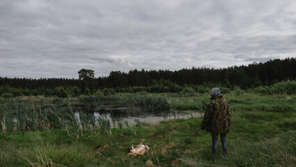 A girl in a camouflage jacket walks a dog on a leash next to a pond where tall grass and reeds grow
