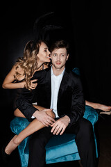 Obraz premium Beautiful woman biting ear of handsome boyfriend in suit sitting in armchair isolated on black