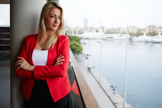 Self confidence concept, attractive businesswoman looking to you while standing at office balcony with beautiful seaport view on background, female executive with crossed arms looking to the camera