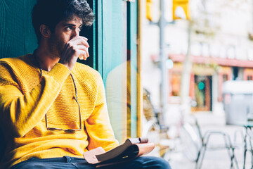 Fototapeta premium Bearded creative young man drinking tasty coffee while looking out of window and thinking on new ideas for creating schedule.Thoughtful male dressed in casual yellow sweater enjoying beverage