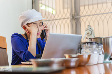 Happy woman baker talking on cell phone with laptop and bakery ingredient on table.