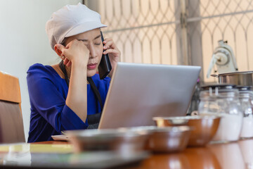 Serious woman baker talking on cell phone with laptop and bakery ingredient on table.