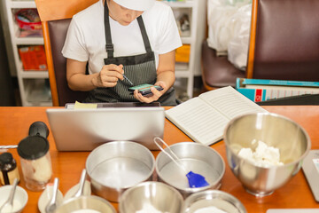 Female baker using cell phone with laptop and bakery ingredient on table.