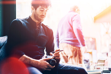 Professional male photographer dressed in black shirt chaecking and making setting on vintage camera to take photos while sitting on windowsill of stylish coworking space during leisure time